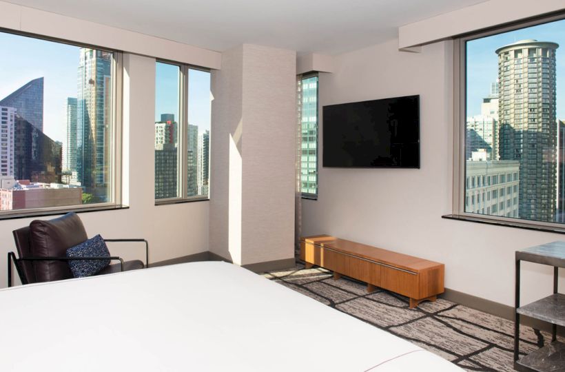 A modern room with a bed, chair, wall-mounted TV, and city views through large windows, featuring skyscrapers outside.