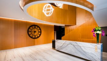 A modern interior with a marble reception desk, wooden paneling, large wall clock, spiral lighting, and flowers on the counter.