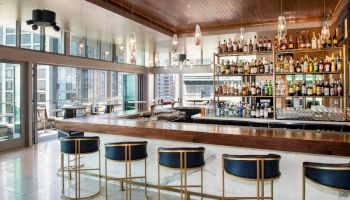 A modern bar interior with high stools, a stocked liquor shelf, and large windows offering city views.