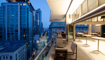 A rooftop terrace overlooking a cityscape at dusk with seating areas and modern high-rise buildings surrounding it.