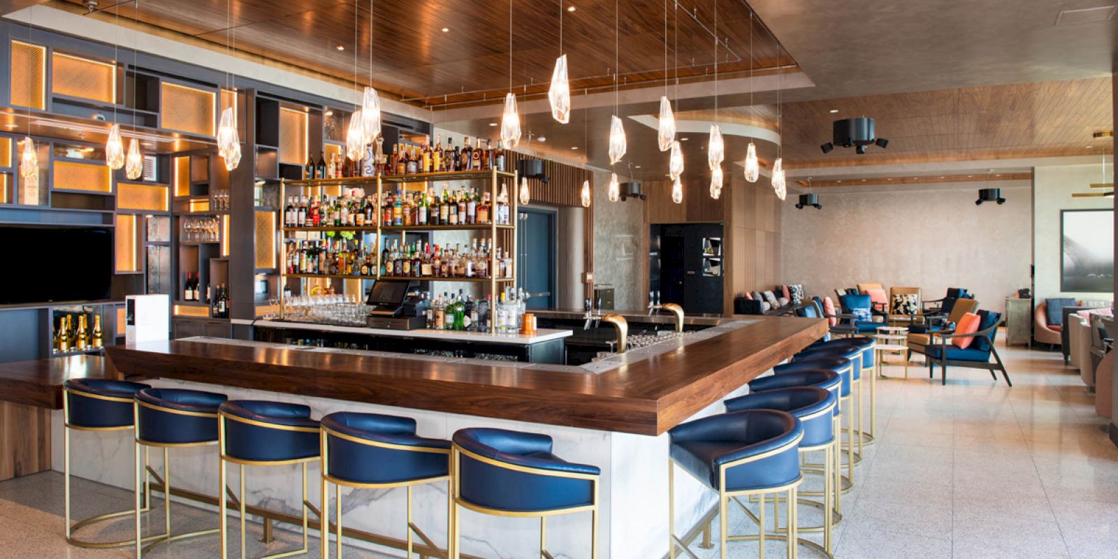 A modern bar interior with blue barstools, a wooden counter, hanging lights, and a well-stocked shelf of bottles, offering a cozy ambiance.