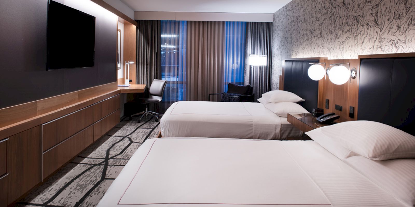 The image shows a modern hotel room with two beds, a desk, chair, TV, and stylish decor. The room is well-lit and neatly arranged.