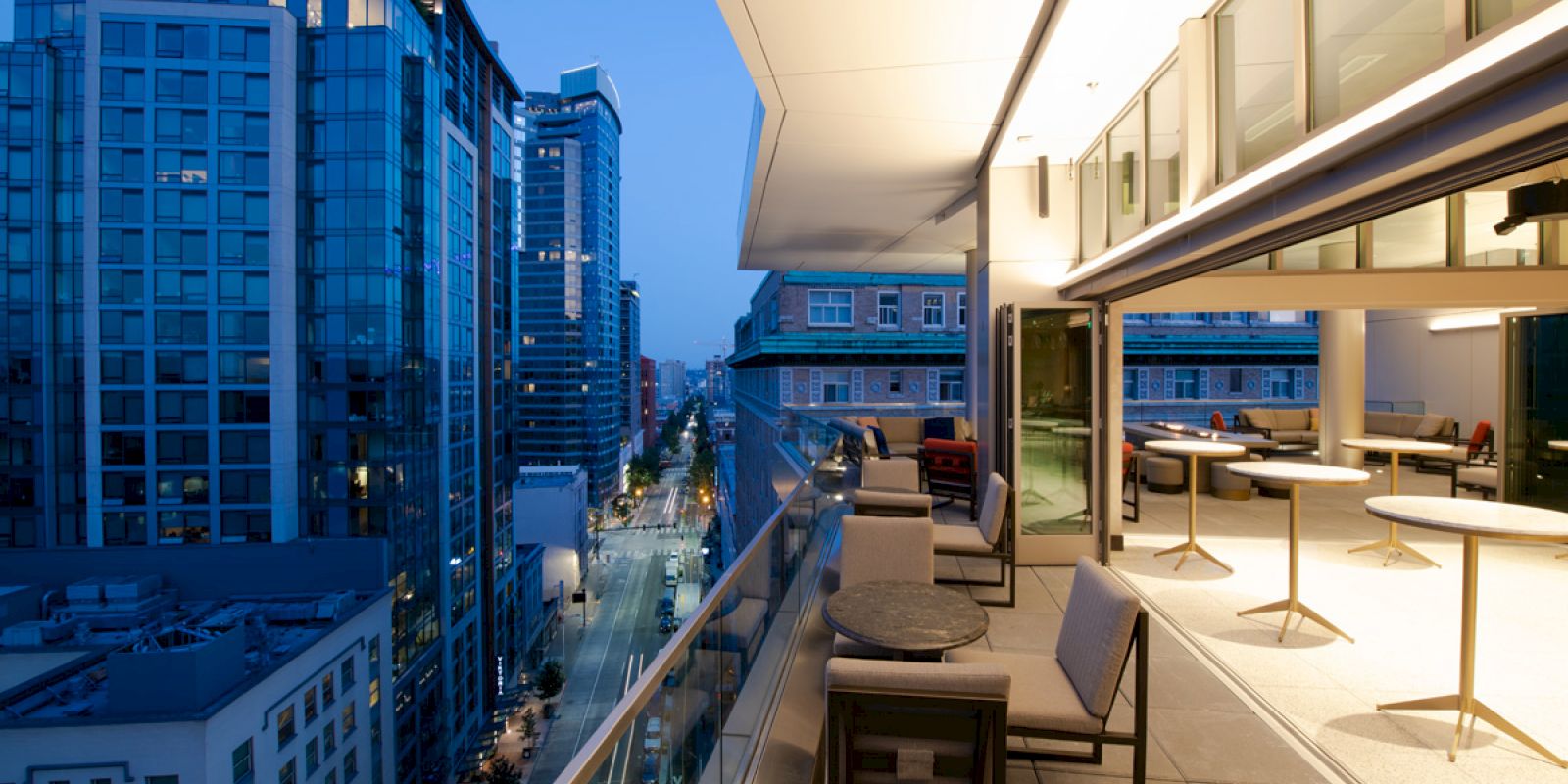 A modern outdoor terrace at dusk with seating and tables overlooks a city street lined with tall office buildings.