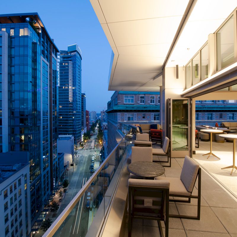 A modern rooftop terrace at dusk with outdoor seating overlooks a city street lined with tall buildings.