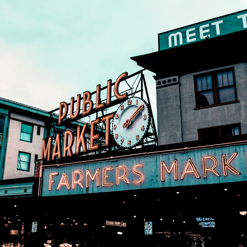 The image shows a neon sign for a "Public Market" and "Farmers Market" with a clock, on a building, under a cloudy sky.