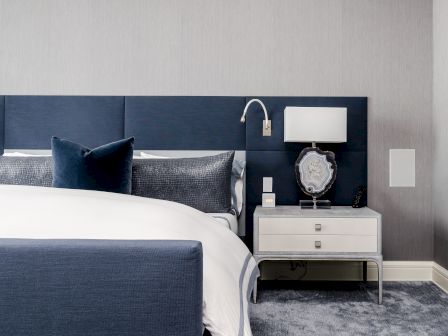 A modern bedroom features a navy headboard, crisp white bedding, nightstand with lamp, decorative stone, and wall-mounted reading light.