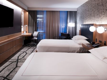 The image shows a modern hotel room with two beds, a wall-mounted TV, a desk, and artistic wallpaper, creating a cozy atmosphere.