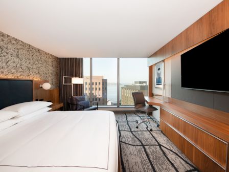 A modern hotel room with a large bed, wall art, desk, chair, and a TV. Large window with a city and water view completes the setting.