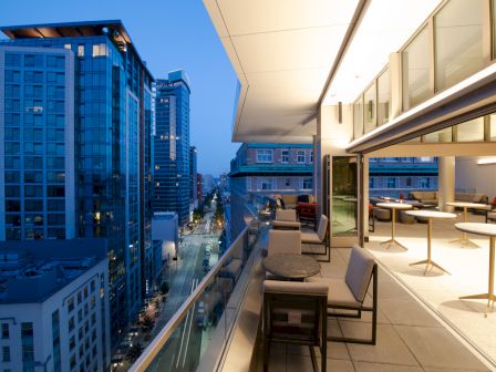 A modern rooftop terrace with seating overlooks a city street lined with tall buildings at dusk, under a clear sky.