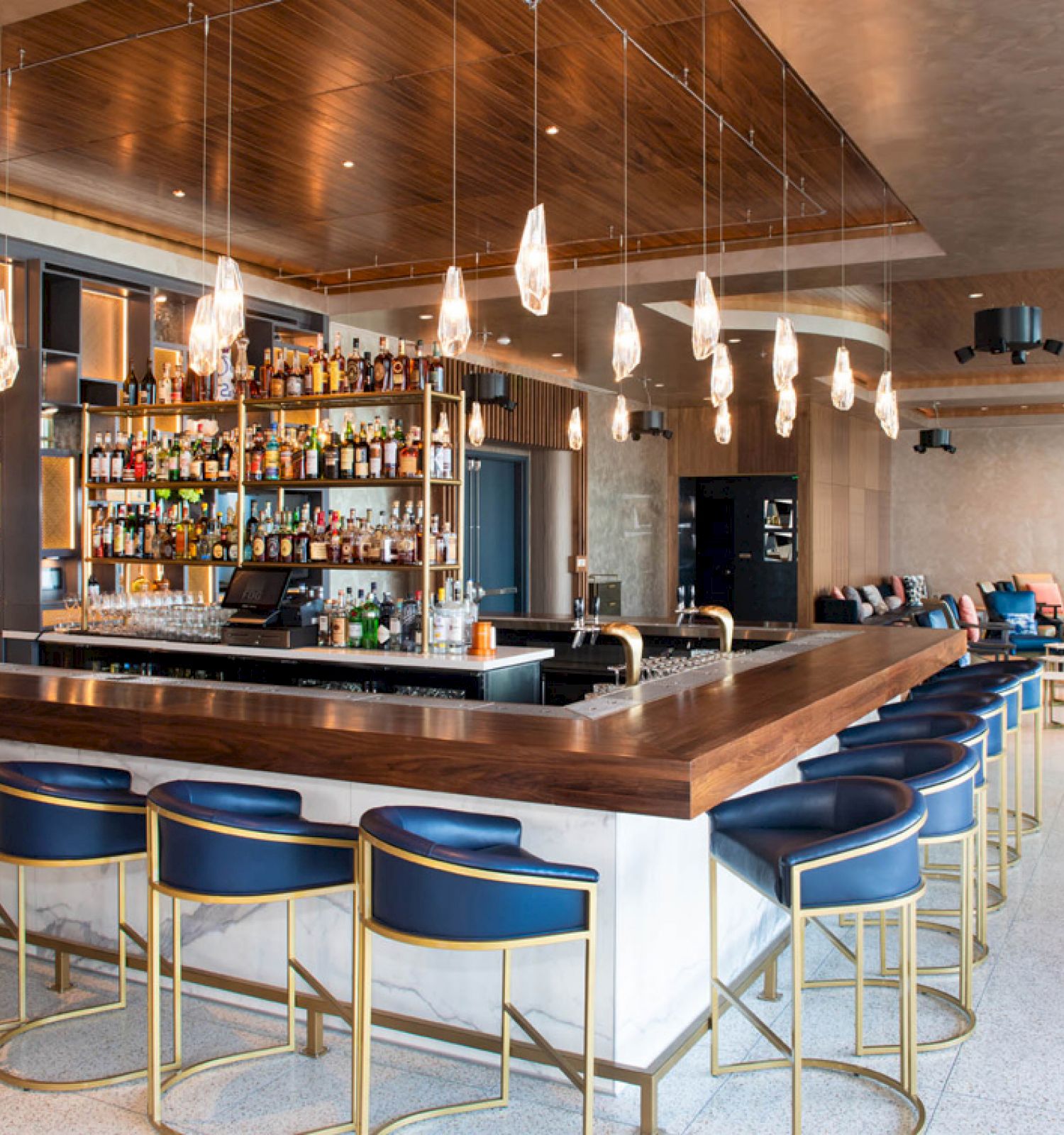 This image shows a modern bar with a central counter, blue stools, hanging lights, and a well-stocked liquor shelf, in a contemporary setting.