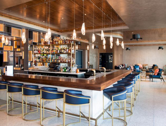 The image shows a modern bar with blue bar stools, wooden accents, hanging lights, and a well-stocked shelf of bottles.