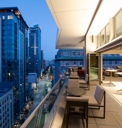 A modern cityscape at dusk shows a high-rise balcony with seating, overlooking a street lined with illuminated buildings and a twilight sky.
