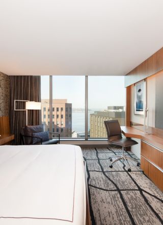 A modern hotel room features a large bed, desk with chair, flat-screen TV, and a window with a cityscape view and water in the background.