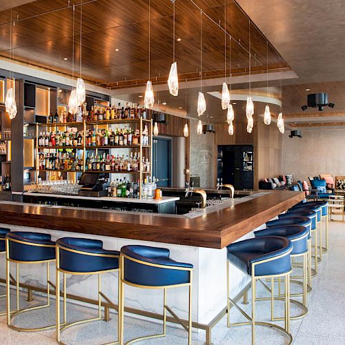 A modern bar with a variety of liquor bottles, stylish seating, pendant lighting, and a seating area in the background is shown.