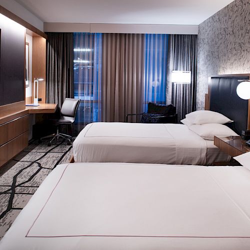 A modern hotel room with two beds, a desk, chair, TV, and ambient lighting creating a cozy atmosphere.
