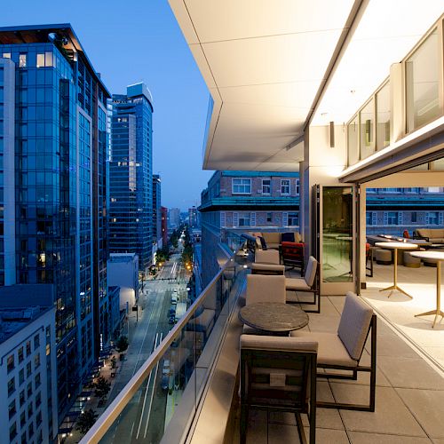 A modern cityscape view from a balcony with seating, tall buildings, and a brightly lit interior space at dusk.