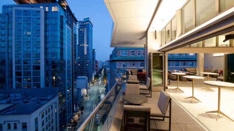 A modern balcony view overlooking a cityscape at dusk, with outdoor seating and indoor tables and chairs visible inside.