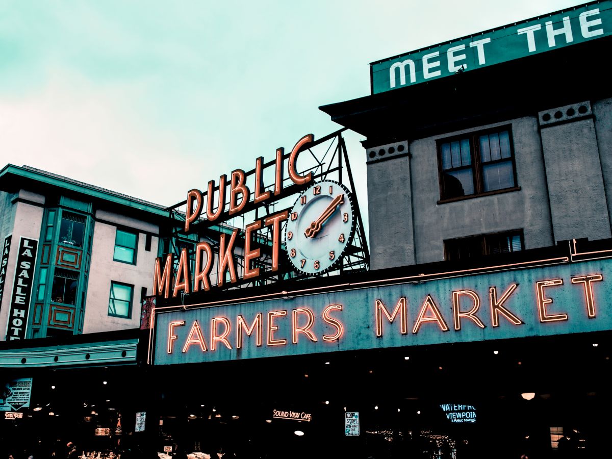 A historic building with signs saying "Public Market" and "Farmers Market," and a clock in an urban setting under a cloudy sky.