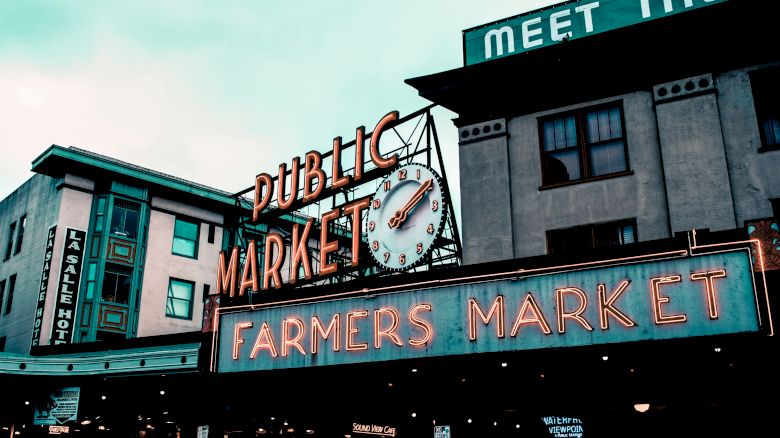 A historic building with signs saying "Public Market" and "Farmers Market," and a clock in an urban setting under a cloudy sky.