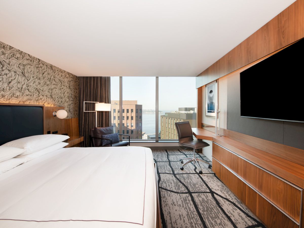 A modern hotel room with a large bed, desk, chair, wall-mounted TV, and a window view of buildings and water.