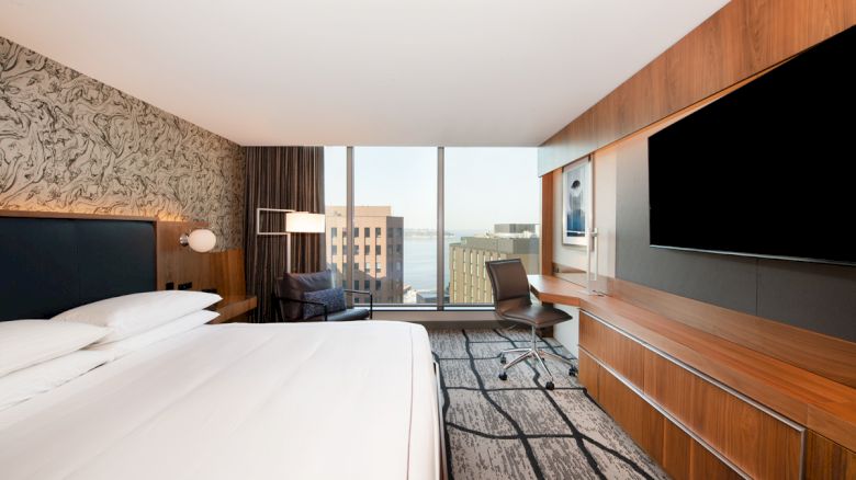 A modern hotel room with a large bed, desk, chair, wall-mounted TV, and a window view of buildings and water.