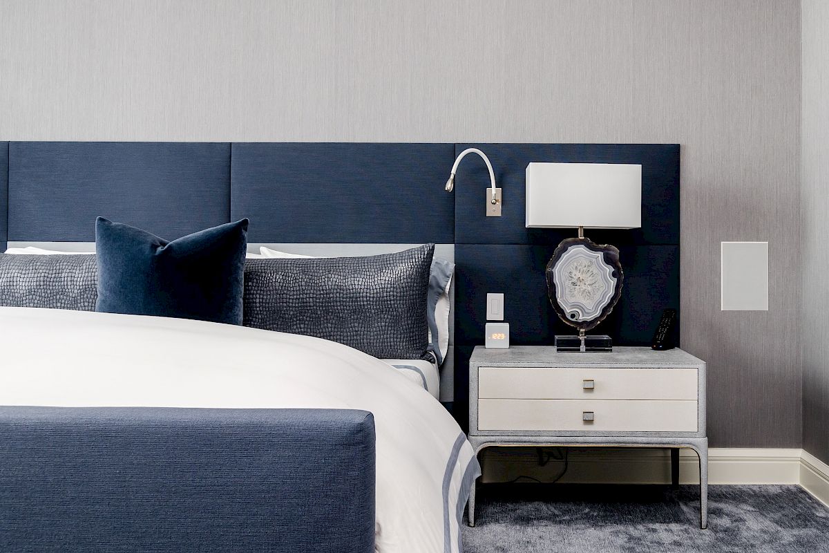 A modern bedroom with a navy-blue bed, gray pillows, white nightstand, stylish lamp, and wall-mounted light fixture.