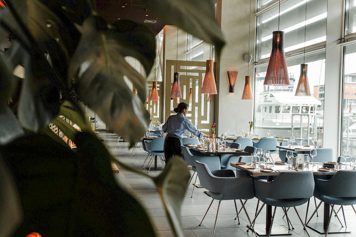 A restaurant interior with modern decor, a person setting tables, stylish pendant lights, and large windows.