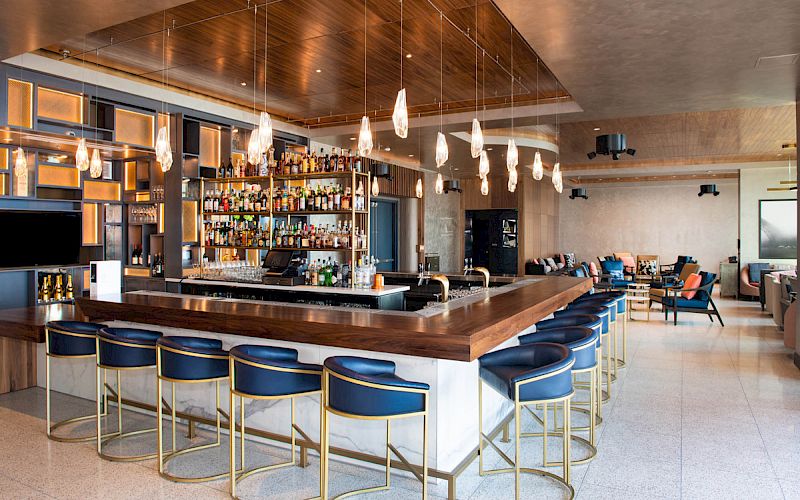 The image shows a modern bar with blue stools, a well-stocked backlit bar, and pendant lights, in a spacious room with seating areas.