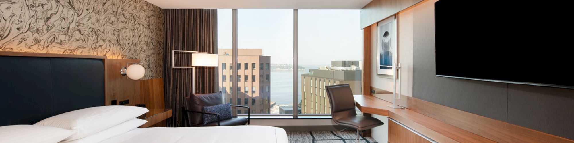 A modern hotel room with a large bed, wall-mounted TV, desk, and large window offering a city and water view.